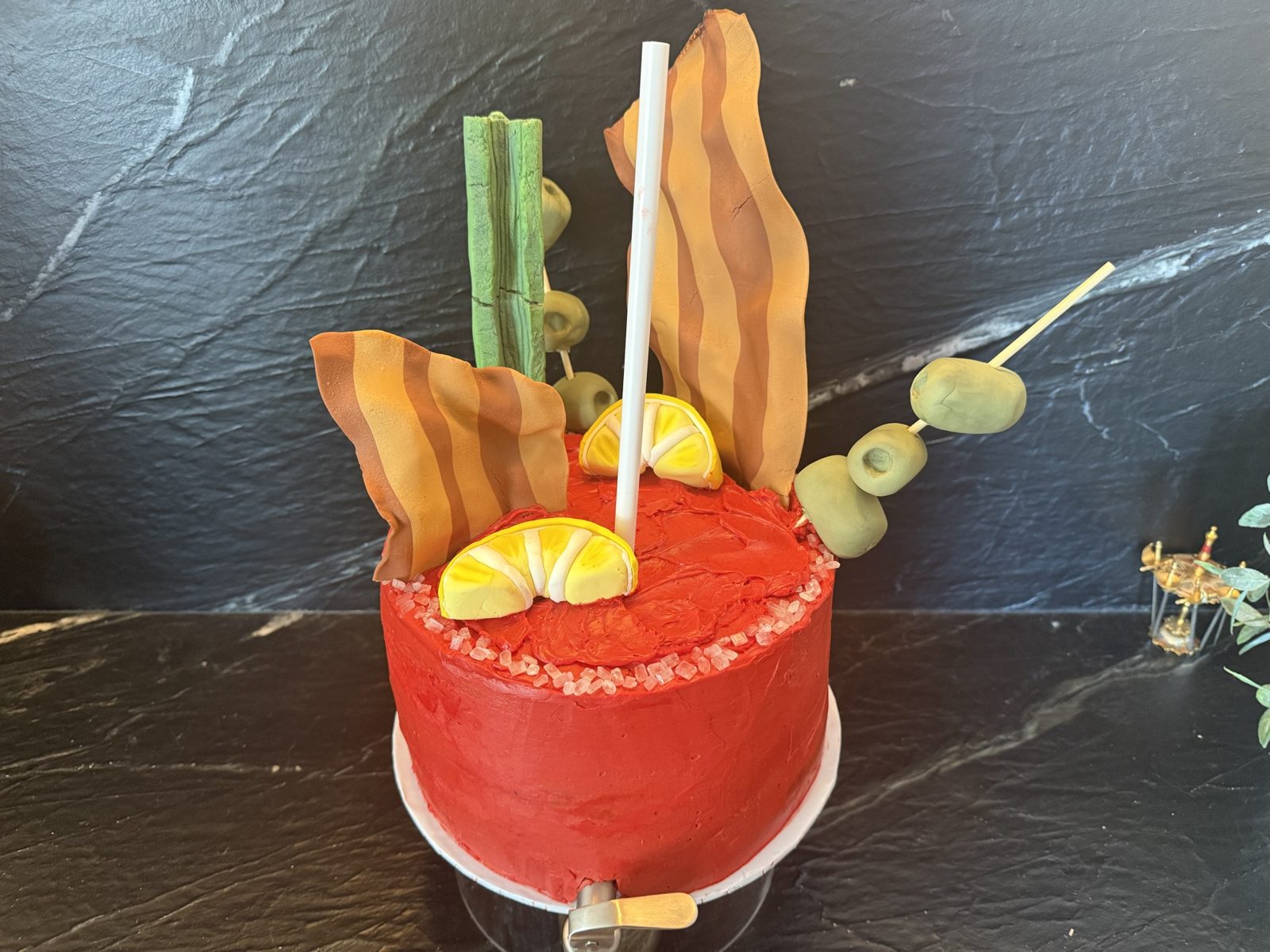 A red buttercream cake made to look like a Bloody Mary, topped with fondant bacon, celery, lemon wedges and an olive skewer